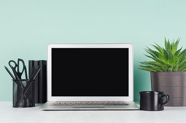 Spring workplace with blank notebook screen, black stationery, books, aloe plant in ribbed pot, coffee cup, in elegant green mint menthe interior on white wood table.