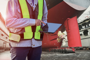 Surveyor, Engineer search on hand holding tablet on ship propeller, rudder red stern ship after maintenance and ship repair floating dry dock in shipyard Thailand.