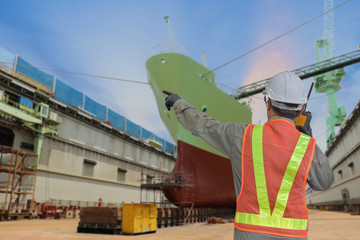 Worker in floating dry dock hand holding walkies talkie finger point on cargo ship moored in...
