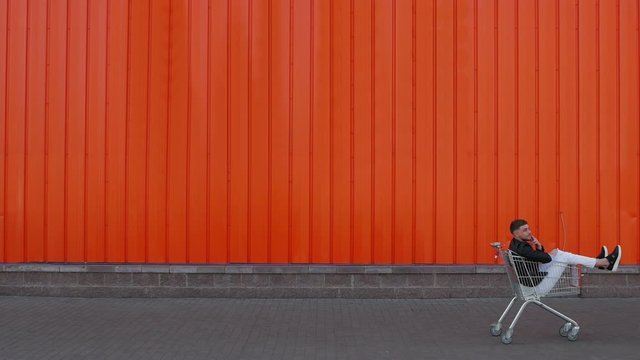 Happy customer man is sitting in a shopping cart and rides along the wall of mall store center outside and imitates smoking. Discounts and sales concept. Funny and amusement moment