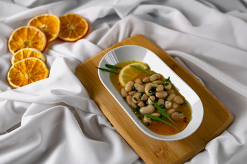 Marinated white beans in their own juice on an oval plate with Thai pineapple sauce and green onions. Decorated with a slice of lemon and dried oranges on a wooden Board with a snow white fabric