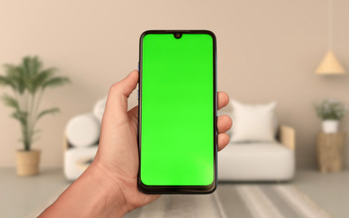 Isolated man hand holding smartphone with Green mock up sreen on living room background.