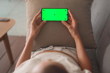 Woman lying down on couch and holding smartphone with green screen in horizontal landscape mode