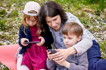 A young woman with a boy and a girl are seated in the forest with mobile phones. Against the...