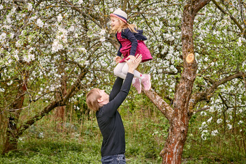 A young father plays with his daughter in a flowering garden. Throws up. Against the background of green grass and flowering trees.