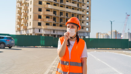 Female construction worker in overalls and medical mask coughing into fist on background of house under construction. Concept of threat of coronavirus epidemic infection.