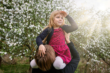 Obraz premium The girl sits on her father’s shoulders in the sun. Against the background of a blooming garden. A happy family.