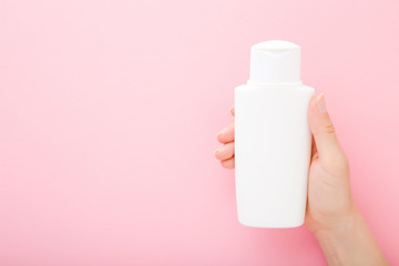 Young woman hand holding and showing white shampoo bottle on light pink table background. Pastel...