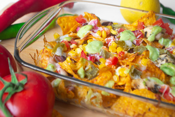 Baked nacho dish in a glass baking pan with corn tortilla chip, pico de gallo salsa, avocado guacamole chipotle, beans, jalapenos and Monterrey jack cheese on a wooden board surrounded by ingredients