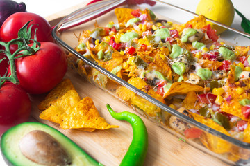 Baked nacho dish in a glass baking pan with corn tortilla chip, pico de gallo salsa, avocado guacamole chipotle, beans, jalapenos and Monterrey jack cheese on a wooden board surrounded by ingredients