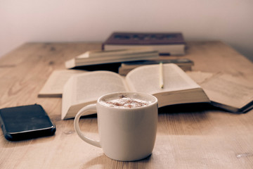 white cup with cappuccino coffee accompanied by a series of books and a cell phone on a rustic wooden table