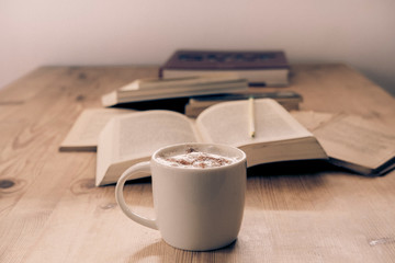 white cup with cappuccino coffee accompanied by a series of books on a rustic wooden table