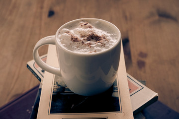 white cup with cappuccino coffee on top of a small tower made up of books inside a hipster room