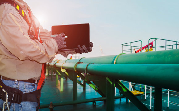 Crew Worker On Oil Tanker Storage Ship And Hand Holding Ipad Wearing Safety Full Harness With Green Pipe Line Connect Existing On Deck.