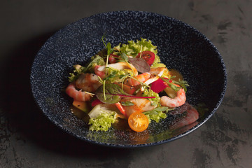 Restaurant menu. Salad with king prawns, squid, herbs and tomatoes. The blue plate. Dark background. Red, orange, green, yellow