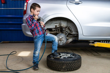 A young boy, a young auto worker, makes a tire change with a pneumatic wrench in the garage of a...