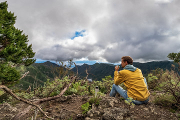 Naklejka premium Young male traveler drinks a hot drink in the mountains. Drinking tea or coffee from thermos in the nature. Dreaming and looking into the distance on cloudy day.