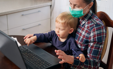 Stay at home mother in a mask working remotely while taking care of her baby. Modern technologies help to keep the job. Quarantine isolation covid-19 concept