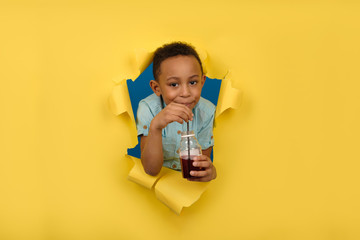 Happy African-American boy in blue shirt is holding juice bottle, drinking red cherry drink from...