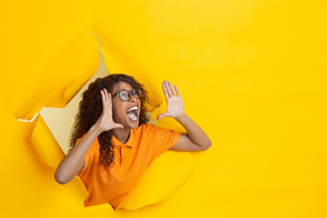 Astonished shouting. Cheerful african-american young woman in torn yellow paper background,...