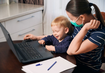 Self isolation to protect your family from covid-19 virus. Mother wearing a mask stays at home working at computer holding her little cute son on her laps. Spending more time together with family