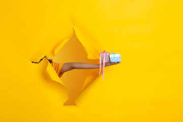 Female hand giving presents, gift, surprise in torn yellow paper hole background. Breaking on, breakthrought. Concept of celebration, shopping, proposal, sales, ad. Copyspace. Promotions discounts