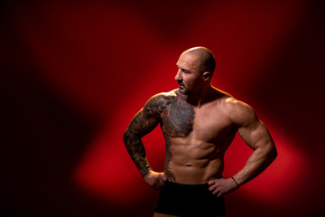 Fototapeta na wymiar A sports man with a tattoo posing on red background. Fitness trainer with naked torso, standing in the studio. Bodybuilder looking away