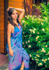 Beautiful young woman posing against a backdrop of greenery and a bungalow with a blue tissue. Elegant girl with scarf and bijouterie enjoying at beach. Freedom and carefree concept, Bali. tanning