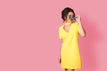 Fototapeta premium Beautiful girl wearing yellow dress and sunglasses posing on pink background in studio. Looking at camera.