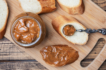 Boiled condensed milk with a glass jar on a wooden background. Sweet paste spread on slices of bread for Breakfast and dessert.