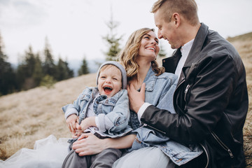 Stylish family in the autumn mountains. A guy in a leather jacket and a young girl in a gray-blue wedding dress with their son sitting on the grass together on the background of forest and landscapes