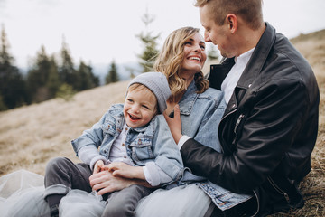 Fototapeta premium Stylish family in the autumn mountains. A guy in a leather jacket and a young girl in a gray-blue wedding dress with their son sitting on the grass together on the background of forest and landscapes