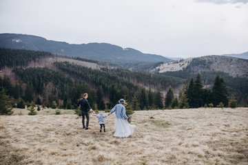 Stylish family in the autumn mountains. A guy in a leather jacket and a young girl in a gray-blue wedding dress with their son walking along the slope in the background of forest and landscapes