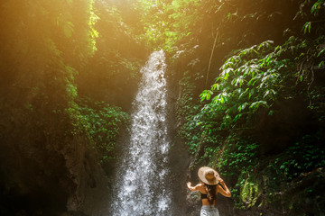 Fototapeta na wymiar Beautiful young woman tourist with straw hat from back. Walks in the tropical jungle and looks at a strong and rapid waterfall illuminated by sunlight. Adventure, tourism, exploration and travel conce