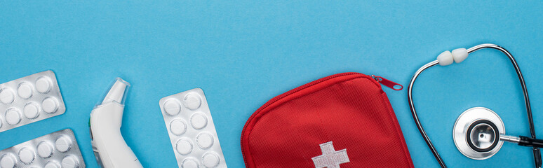 top view of pills in blister packs, stethoscope, first aid kit and ear thermometer on blue background, horizontal image