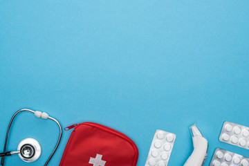 top view of pills in blister packs, stethoscope, first aid kit and ear thermometer on blue background