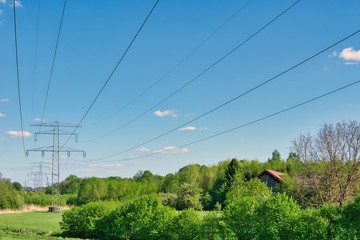 Obraz premium The power cords over a green scenery with trees and a house and under a blue sky with clouds