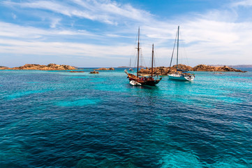 A view of an ancient and a modern sailboat moored in a wonderful and colorful bay of the Mediterranean sea with some rocks in the background on a sunny day with some clouds in summer, Sardinia Italy
