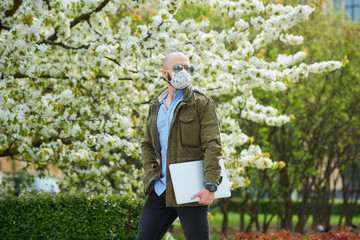 Fototapeta premium A bald man with a beard in a medical face mask to avoid the spread coronavirus walks with a laptop in the park. A guy wears n95 face mask and a pilot sunglasses in the garden between flowering trees.