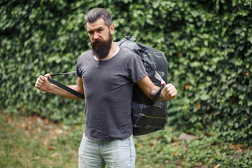 Brutal bearded male courier in shorts and a T-shirt with a big gray bag on his shoulders in a green park. homeless not shaved man with a big gray bag.