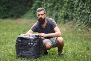 Naklejka na ściany i meble Brutal bearded male courier in shorts and a T-shirt with a big gray bag is resting in a green park. homeless man not shaved with things in nature.