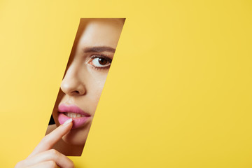 Woman looking at camera across quadrangular hole in yellow paper and touching pink lip