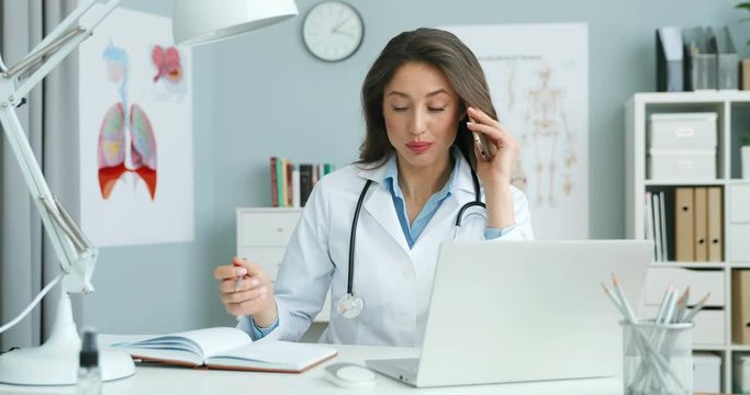 Female doctor sitting at desk with laptop computer and talking on mobile phone and writing down appointment in notebook. Woman medic speaking on cellphone and noting consultation time in planner.