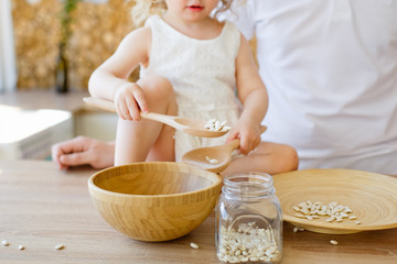 A little girl is sitting on a table in a bright Scandinavian-style kitchen. Playing with dishes, cereals, beans. The concept of children's development Montessori.