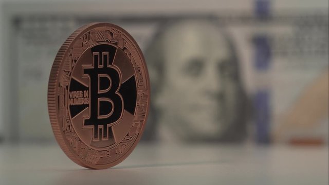 Cripto coin with Bitcoin symbol isolated on foreground. Coin rotate. 100 US dollars is ont the background with nice blur. Virtual money of the future. Close up shot.