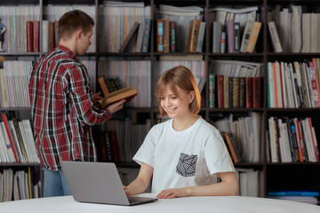 Cute boy and girl students working on a project in the city library, looking for books and typing notes on a laptop.