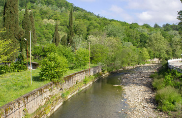 Spring landscape. A mountain meandering river enclosed in stone fences. Shallow, with a bare stone bottom on the side. Green plants, shrubs, and trees. Sunny weather.