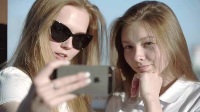 Two young smiling hipster blond women in summer white t-shirt clothes. Girls taking selfie self portrait photos or video on smartphone. Female showing positive face emotions