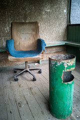 An old dirty vintage armchair covered with a blue rag and a green rusty metal urn for cigarette butts and rubbish against a rough concrete wall.