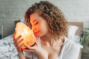 attractive dreamy girl with closed eyes holding Himalayan salt lamp in bedroom